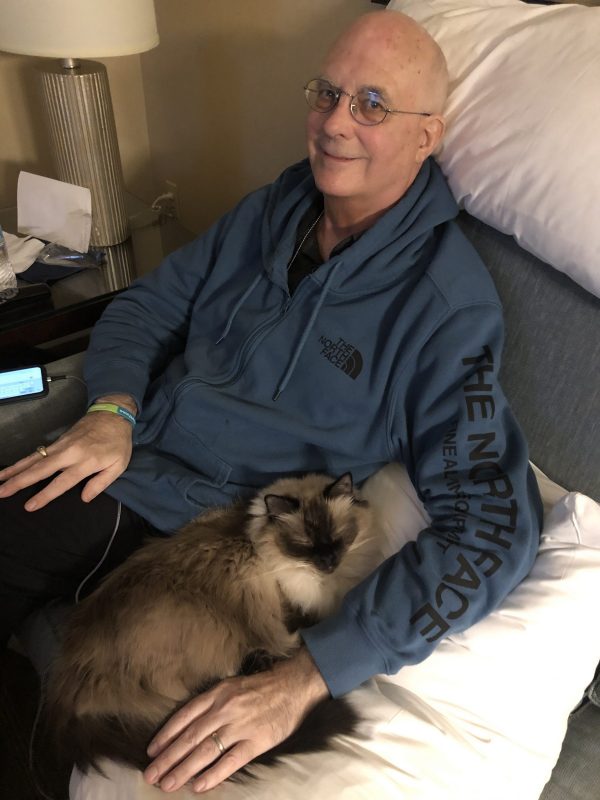 care partner | Pulmonary Fibrosis News | Wearing a blue hoodie, Kevin smiles as he sits in a gray chair with pillows while being comforted by a big, furry cat named Raj