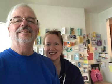 caregiver | Pulmonary Fibrosis News | Samuel Kirton and his wife, Susan, stand in front of a wall of greeting cards that hang in their post-lung transplant apartment