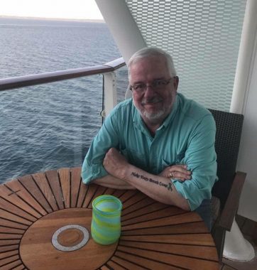 rare disease advocacy | Pulmonary Fibrosis News | A snapshot shows Sam smiling at a table on a deck overlooking a body of water. A tattoo on his forearm is visible.