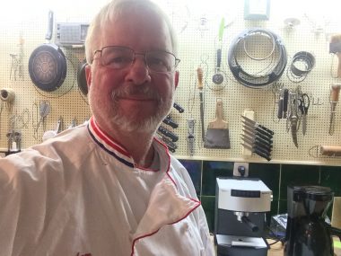 self-care | Pulmonary Fibrosis News | Sam wears a white chef's apron and stands in front of a wall of cooking utensils at Julia Child's home in Plascassier, France.