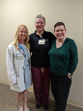 Three nurse practitioners stand in a hallway and smile with their arms around one another. The nurse on the left has long blonde hair and is wearing a blue striped dress, white coat, and stethoscope. The nurse in the middle has glasses and is wearing a black collared shirt, burgundy pants, and an ID. The nurse on the right has brown hair and is wearing a long-sleeve green shirt, black pants, and a smart watch.