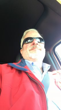 This photo is angled upward to capture the face of a man with gray hair, mustache, and beard. He's wearing sunglasses and a red jacket with a light shirt and is strapped into a seat belt in a car.