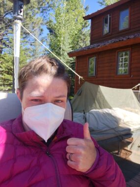 A woman wearing a magenta down jacket and a white face mask gives a thumbs up while sitting outside on her parents' deck. Part of a wooden home is visible in the background, along with tall trees. A tent is set up on the deck to accommodate the woman while she recovers from COVID-19.