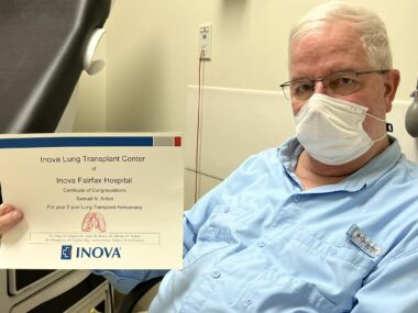 A man with white hair and glasses holds up a certificate from the Inova Lung Transplant Center of Inova Fairfax Hospital that recognizes the second anniversary of his bilateral lung transplant. The man is wearing a long-sleeve, light blue Columbia shirt and a white disposable face mask. He appears to be sitting in a doctor's office.