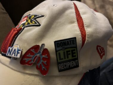 A close-up of a baseball cap with various pins, including a pin depicting a set of lungs and a pin demonstrating that the wearer is the recipient of donated organs. The photo is really dark and the specific items are not entirely visible. 