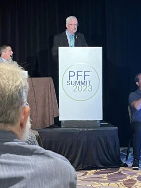 A man with gray hair, a goatee, and glasses stands at a silver lectern that reads "PFF Summit 2023" in a circle at the front. He wears a dark sport coat with a green tie; we see a man at the front table at his left and, on the same side, shoulders of audience members. Another man, seen in part, is seated in a chair at his right. A black curtain is the backdrop, and the floor has a purple and yellow design.