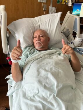An older man lies in a hospital bed surrounded by monitors and machines. He's wearing a hospital gown and bracelet, and though his expression is solemn, he's giving the camera two thumbs up.