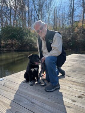 A man kneels next to a puppy on a wooden pier near his home in Virginia. He's wearing a long-sleeve khaki shirt, black vest, blue jeans, and sunglasses. The puppy is a mostly black Aussiedor with several white patches on his chest and paws. It's a beautiful, sunny day, and both the man and the dog look very happy.