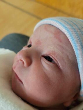 A close-up photo shows a newborn baby wrapped in a white blanket and wearing a blue and white striped hat.