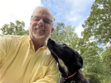 A man with white hair, a goatee, and glasses takes a low-angle selfie of himself and his dog. The man appears to be sitting or squatting outside, while the dog is looking up at him lovingly. In the background we can see trees and a partly cloudy sky.