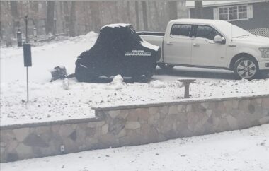 A gray stone retaining wall separates one area of light snow from another, raised area. We see a white truck and a black object in the raised area, with what appears to be a house in the distance.