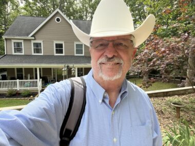 A man with glasses, a gray mustache, and a goatee stands before a two-story house surrounded by trees, some flowering. He wears a blue, long-sleeve Oxford cloth shirt and a white Stetson hat, with a visible shoulder bag harness.