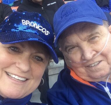 A couple smiles for a close-up selfie in a football stadium. They are both wearing blue baseball caps and blue shirts, and the man has a nasal cannula for his supplemental oxygen.
