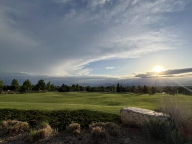 The sun sets over a large grassy field lined by trees in the distance.