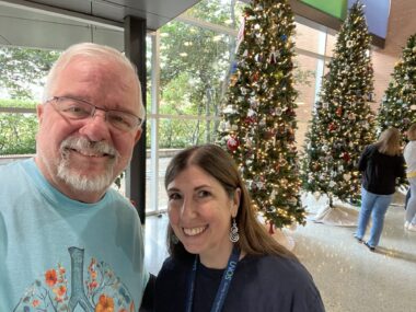Two people pose for a photo in a spacious lobby with large windows. Three tall decorated holiday trees are visible behind them.